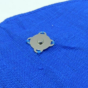 May include: A silver magnetic snap closure attached to a piece of bright blue fabric. The snap has a circular center and a decorative, four-lobed outer design. The fabric appears to be a woven material.