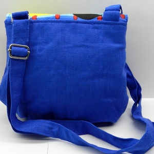 May include: A small, square blue crossbody bag with a matching adjustable strap. The bag features a colorful patterned top edge with red stitching. The strap is attached with a silver-colored metal buckle.
