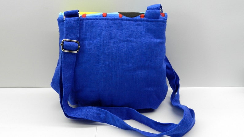 May include: A small, square blue crossbody bag with a matching adjustable strap. The bag features a colorful patterned top edge with red stitching. The strap is attached with a silver-colored metal buckle.