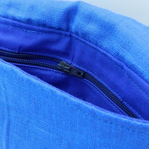 May include: A close-up of a blue linen bag with a flap. The bag's interior is a darker shade of blue, with a zippered pocket. The bag is open, revealing the inside. The bag has a strap.