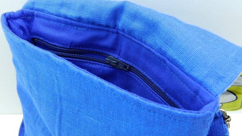 May include: A close-up of a blue linen bag with a flap. The bag's interior is a darker shade of blue, with a zippered pocket. The bag is open, revealing the inside. The bag has a strap.