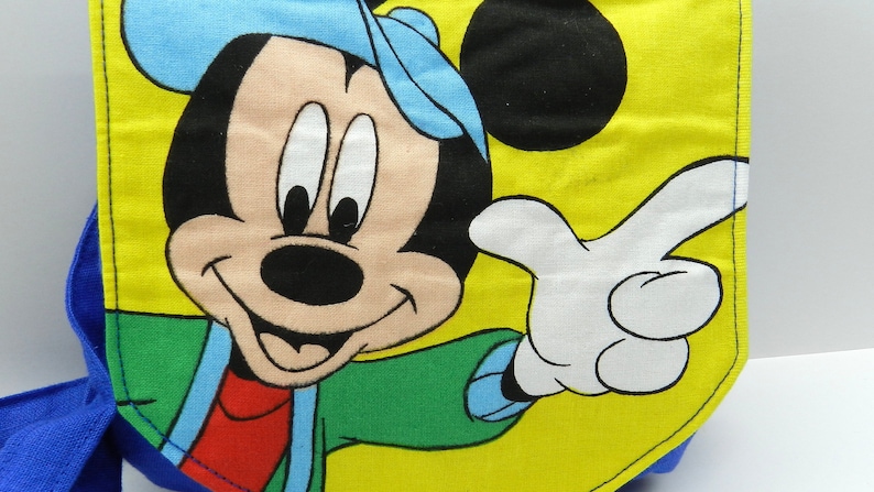 May include: A yellow and blue backpack featuring a large graphic of Mickey Mouse. The cartoon character is wearing a blue hat and green jacket, with a white gloved hand pointing. The backpack has blue straps.