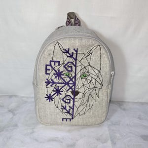 Op de afbeelding: Een grijze rugzak met een geometrisch wolfontwerp en paarse sneeuwvlokborduurwerk. De wolf heeft groene ogen. De rugzak heeft een ritssluiting en een handvat met patroon.