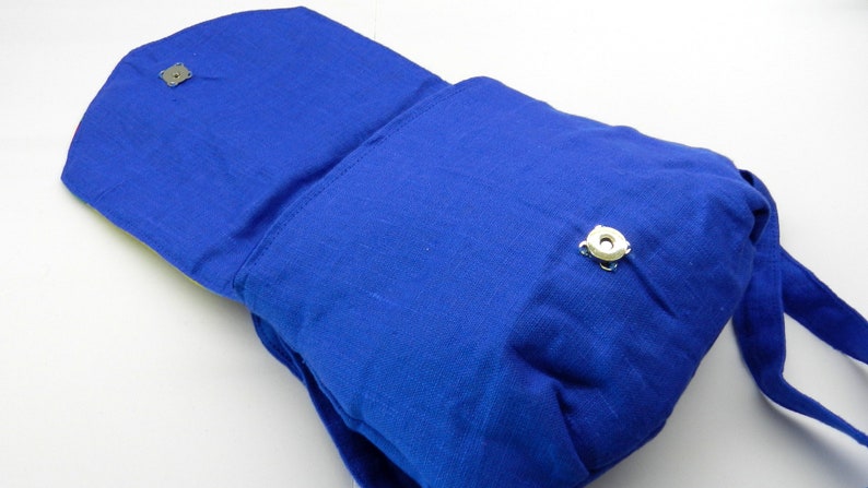 May include: A vibrant blue fabric bag with a flap closure and a magnetic snap. The bag has a rectangular shape with rounded edges and a strap. The interior is a lighter color, and the bag appears to be made of linen or a similar material.