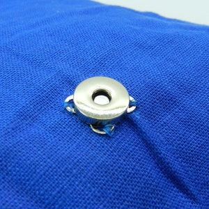 May include: A close-up of a silver snap fastener attached to a vibrant blue fabric. The snap is circular with a central hole and is secured with small loops of blue thread. The fabric has a textured appearance.