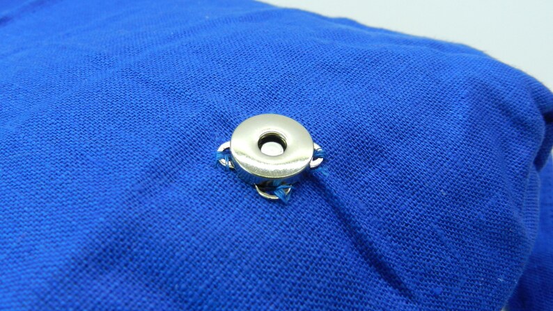 May include: A close-up of a silver snap fastener attached to a vibrant blue fabric. The snap is circular with a central hole and is secured with small loops of blue thread. The fabric has a textured appearance.
