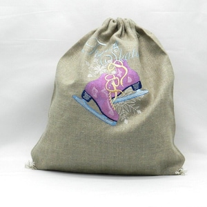 Könnte beinhalten: Ein beigefarbener Jutebeutel mit Kordelzug und einer Stickerei, die lilafarbene Schlittschuhe mit gelben Schnürsenkeln und hellblauen Kufen zeigt. Das Motiv enthält weiße Schneeflocken und den Schriftzug Ice Skates in hellblauer Schreibschrift.