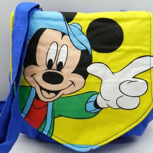 May include: A blue and yellow shoulder bag featuring a large graphic of Mickey Mouse. The bag has a blue strap and trim, with Mickey Mouse in a blue hat, green jacket, and red shirt. The background is yellow.