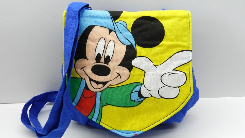 May include: A blue and yellow shoulder bag featuring a large graphic of Mickey Mouse. The bag has a blue strap and trim, with Mickey Mouse in a blue hat, green jacket, and red shirt. The background is yellow.