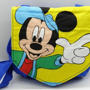 May include: A blue and yellow fabric bag featuring a smiling Mickey Mouse graphic. The bag has a blue strap and a yellow background with Mickey Mouse's face and pointing hand. The bag is likely for children.