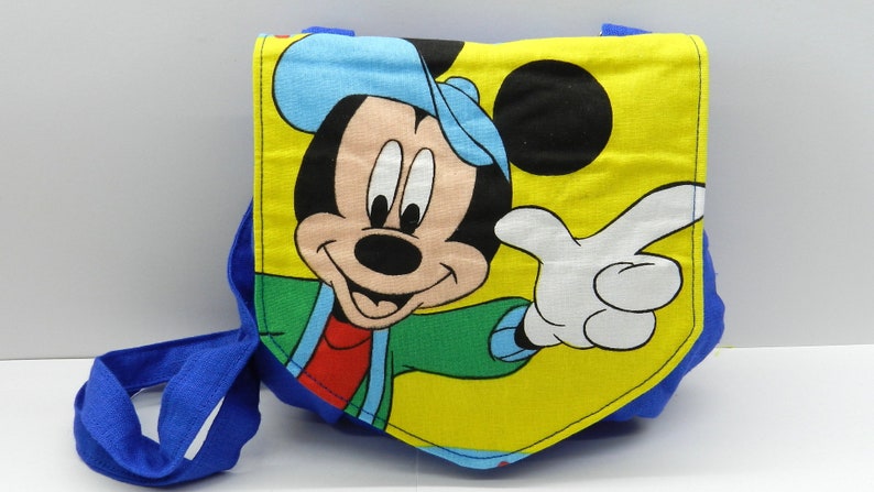 May include: A blue and yellow fabric bag featuring a smiling Mickey Mouse graphic. The bag has a blue strap and a yellow background with Mickey Mouse's face and pointing hand. The bag is likely for children.