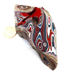 May include: A unique, multi-colored stone with a layered, swirled pattern. The stone features red, white, blue, and black hues. A small, round, off-white object is next to the stone. The stone has a natural, organic shape.