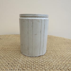 Può includere: Un contenitore cilindrico in ceramica bianco sporco con scanalature verticali. Il vaso in stile antico ha una superficie leggermente strutturata e un bordo spesso. Si trova su una superficie beige intrecciata, suggerendo un'estetica rustica o da fattoria.