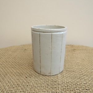 Può includere: Un contenitore cilindrico in ceramica bianco sporco con scanalature verticali. Il contenitore ha una superficie leggermente strutturata e una parte superiore arrotondata. Si trova su una superficie tessuta, color beige. Il contenitore è alto circa 15 cm.