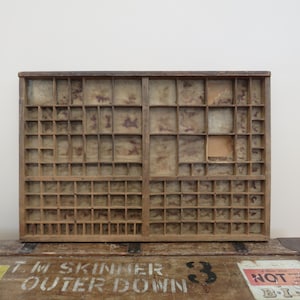 May include: A rectangular wooden letterpress type case featuring numerous small, varied compartments for organising metal type. The wood has a weathered, light brown finish with visible grain and signs of age throughout the interior dividers.