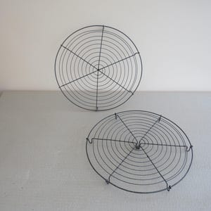 Pode incluir: Duas grelhas redondas de arame cinza escuro. Cada grelha apresenta um design circular concêntrico com suportes radiais. As grelhas são colocadas sobre uma superfície cinza claro contra um fundo branco. Ideal para assar e arrefecer.