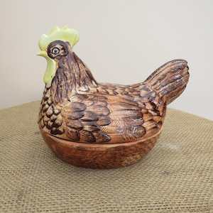 May include: A ceramic chicken-shaped lidded container featuring a brown and tan feathered body with a pale yellow comb and wattle. The base is designed to resemble a textured nest, and the top lid forms the upper body and head of the chicken.