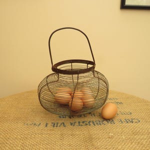 May include: A round, vintage-style wire egg basket with a dark, weathered metal finish and a single arched handle. The basket contains several light brown eggs, and one additional brown egg rests on the surface next to the wire container.
