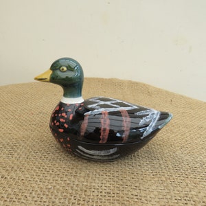 May include: A ceramic duck-shaped container with a glossy finish. The duck has a green head, yellow beak, and a black body with red and white accents. The container is likely used for decorative or storage purposes.