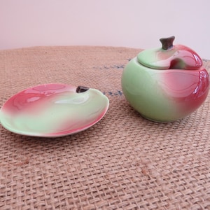 Peut inclure: Un récipient en céramique en forme de pomme avec couvercle et une petite coupelle assortie. Les deux pièces présentent une finition lisse et brillante avec un dégradé de vert tendre et de rouge vif. Le couvercle comporte une tige brune.