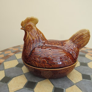 May include: A brown glazed ceramic chicken-shaped lidded dish. The top piece is moulded in the form of a hen with detailed feather textures, a comb, and a wattle. The base is a matching brown bowl with a textured exterior, forming a complete container.