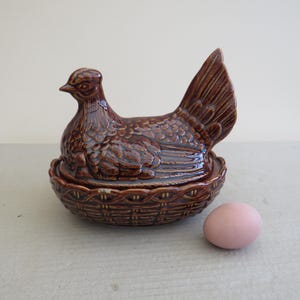 May include: A brown ceramic hen-shaped container with a woven basket base. The hen has detailed feathering and a raised tail. A pink egg rests beside the container. This decorative piece is suitable for kitchen decor.
