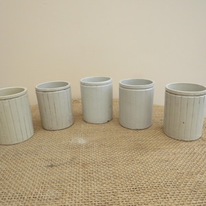 Può includere: Cinque contenitori in ceramica bianco sporco con dettagli a coste verticali. I vasi cilindrici sono disposti su una superficie strutturata marrone chiaro. I contenitori hanno un design semplice e minimalista.