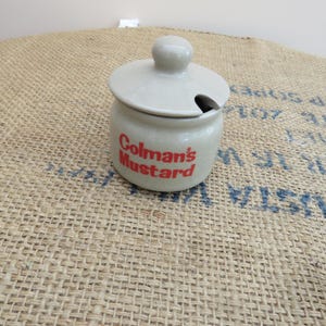 Può includere: Un piccolo vasetto di senape in ceramica bianco sporco con coperchio e fessura per cucchiaio. Le parole "Colman's Mustard" sono stampate in rosso sul davanti. Il vasetto è su una superficie di juta.