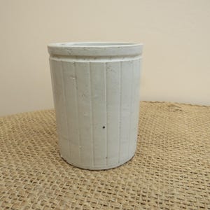 Può includere: Un vaso cilindrico in ceramica bianco sporco con scanalature verticali. Il vaso ha una superficie leggermente strutturata e un bordo arrotondato. È appoggiato su una superficie tessuta, color beige, su uno sfondo neutro. Questo vaso decorativo potrebbe essere usato come vaso o fioriera.