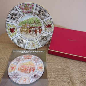 Puede incluir: Un plato decorativo que conmemora el aniversario de la coronación, con una ilustración detallada de una procesión real. El plato tiene un fondo blanco, un borde dorado y patrones decorativos. También hay una caja roja con ribete dorado y el texto "Caverswall".