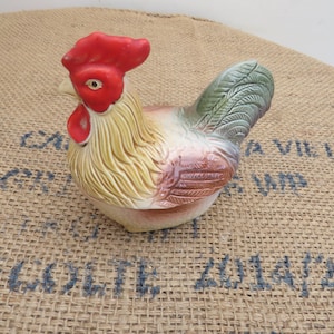 May include: A ceramic rooster figurine with a red comb and wattle, yellow and white body, and green and brown tail feathers. The rooster is sitting on a burlap surface.