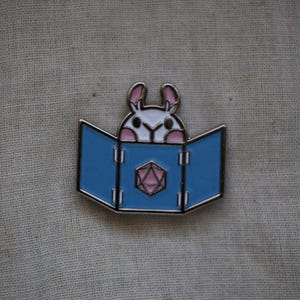 May include: Enamel pin depicting a white bunny peeking over a blue book with a pink and silver geometric design. The pin has a silver border, set against a neutral fabric background.