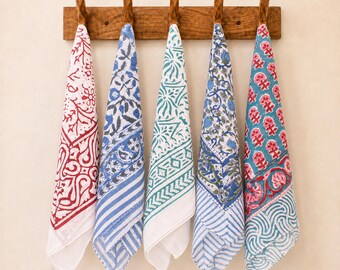 May include: Five rectangular cotton kitchen towels hanging from a wooden rack with five hooks. Each towel features a unique block-printed pattern in colors including red, blue, teal, and green on a white background, showcasing floral and geometric motifs.