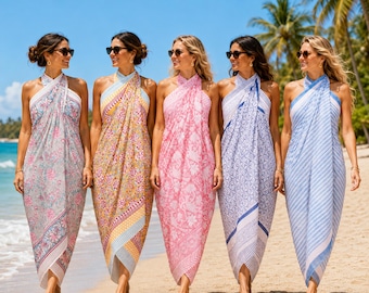 May include: Five sleeveless halter-neck sarong dresses in various patterns and colors, including floral, geometric, and paisley prints in shades of blue, yellow, pink, and white, worn in a line on a sandy beach under a clear blue sky.