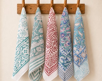 May include: Five rectangular cotton tea towels hanging from a wooden wall rack. Each towel features a distinct block-printed pattern in colors including teal, red, blue, and white, showcasing floral, geometric, and abstract line designs.