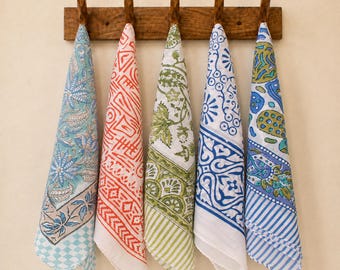 May include: Five rectangular cotton napkins with distinct block-printed patterns in blue, orange, green, and white, hanging from a wooden wall rack with five hooks. Each napkin features a unique combination of floral, geometric, and striped motifs.