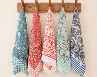 May include: Five rectangular cotton kitchen towels hanging from a wooden wall rack. Each towel features a unique block-printed floral pattern in distinct colorways: teal, coral, white and red, light green, and blue, with checkered or striped borders.