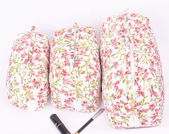 May include: Three white cosmetic bags with a pink floral pattern and gold zippers. The bags are different sizes and have a tassel on the zipper pull. A lipstick and eyeliner pencil are in front of the bags.