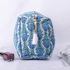 May include: A blue and white quilted makeup bag with a floral print and a white tassel. The bag has a gold zipper and a white tassel.