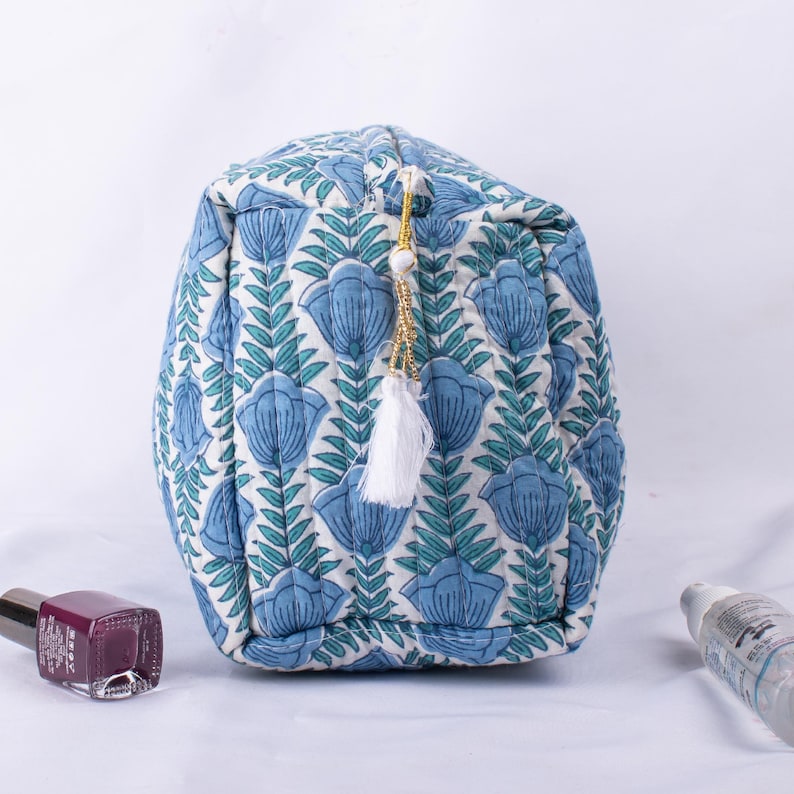 May include: A blue and white quilted makeup bag with a floral print and a white tassel. The bag has a gold zipper and a white tassel.
