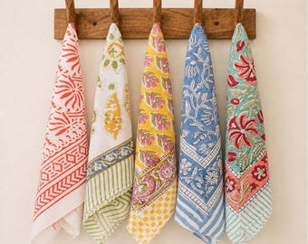 May include: Five rectangular cotton kitchen towels with block-printed floral and geometric patterns in red, yellow, green, and blue, hanging from a wooden wall rack with five hooks. Each towel features a unique color palette and border design.