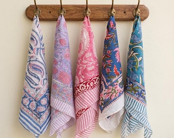 May include: Five square cotton napkins hanging from a wooden rack with brass hooks. Each napkin features a distinct block-printed floral and paisley pattern in various color combinations including blue, pink, lavender, and white with striped borders.