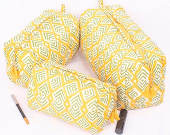 May include: Three yellow and white cosmetic bags with a green geometric pattern. The bags have yellow zippers and tassels. A black lipstick tube and a brown eyeliner pencil are next to the bags.