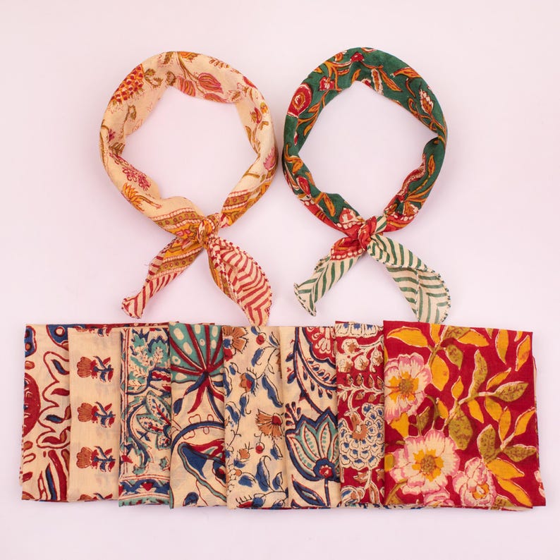 May include: Two tied fabric headbands featuring floral and botanical block print patterns in cream, red, green, and blue, positioned above a row of eight folded fabric squares displaying various colorful floral and leaf motifs in matching prints.