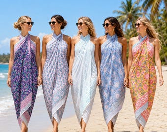 May include: Five halter-neck sarong dresses featuring intricate floral and paisley patterns in purple, light blue, white, and yellow. Each dress has a wrap-style silhouette with a tie at the neck, worn by individuals on a sandy beach.