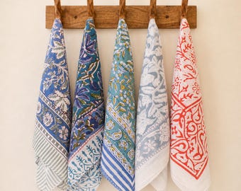 May include: Five rectangular cotton kitchen towels hanging from a wooden rack with five hooks. Each towel features a distinct block-printed pattern in blue, green, grey, and red, showcasing floral, paisley, and geometric motifs on white fabric.