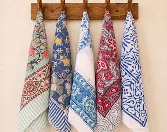 May include: Five rectangular cotton napkins hanging from a wooden wall rack. Each napkin features a distinct block-printed pattern in various color combinations including mint green, navy blue, white, red, and grey with floral and geometric motifs.