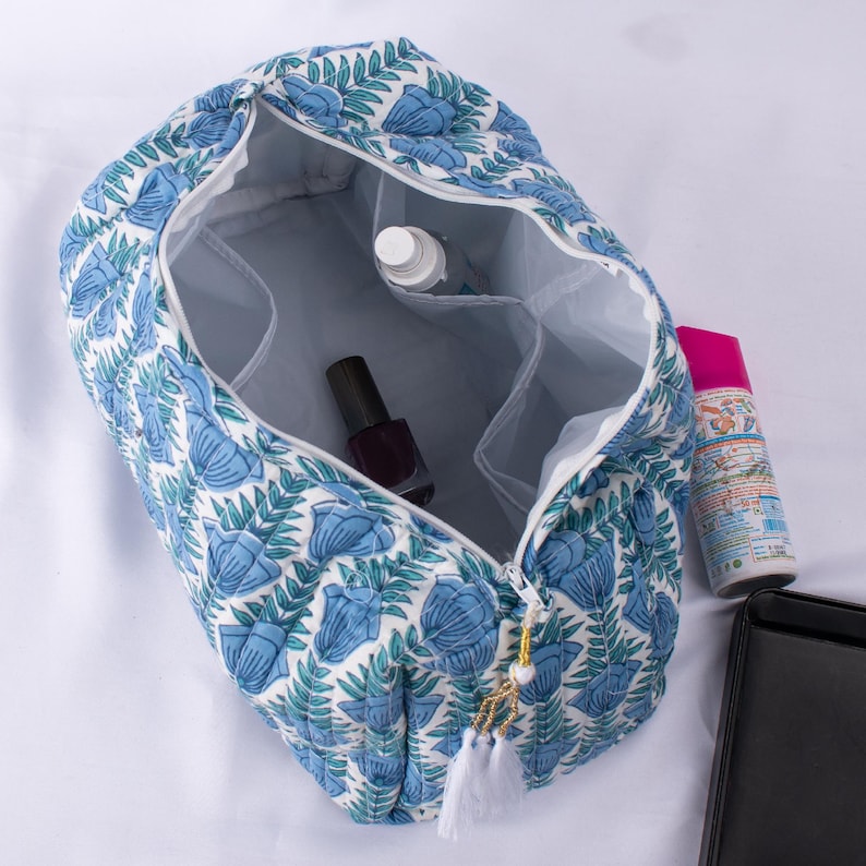 May include: A white quilted makeup bag with a blue floral print. The bag is open and has a zipper closure. The bag has a white tassel.