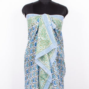 May include: A sarong featuring a white base with a repeating floral pattern in shades of blue and green. The fabric includes a prominent border with blue and white vertical stripes that runs along the top edge and down the centre front drape.