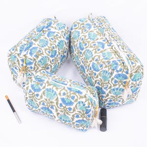 May include: Three white cosmetic bags with a blue floral pattern and green leaves. The bags have a cylindrical shape with a zipper closure and a tassel. A black eyeliner pencil and a black lipstick tube are next to the bags.